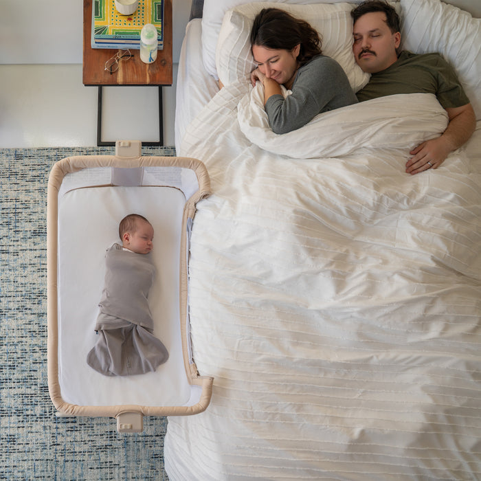 A baby sleeps soundly on HALO 2-in-1 Bedside Bassinet Sleeper Fitted Sheets (100% Organic Cotton, 2 Pack) in a gray blanket next to two adults resting under a white blanket. A nightstand with books and a baby bottle is beside them.