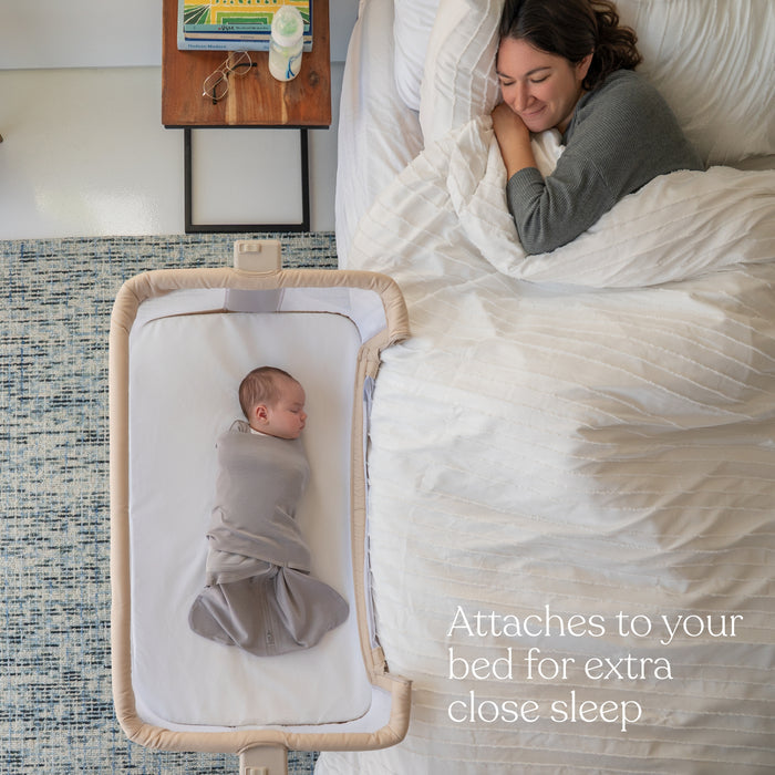 A smiling woman lies in bed under a white comforter, watching her baby swaddled in a HALO SleepSack and resting in the HALO Bedside Sleeper Bassinet Bundle. Text: Attaches to your bed for extra close sleep.
