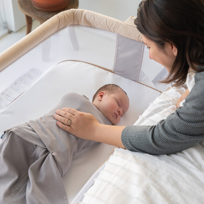 A woman lovingly watches over her sleeping baby, swaddled and lying in a cozy crib fitted with HALO 2-in-1 Bedside Bassinet Sleeper Fitted Sheets, made from 100% organic cotton. Natural light brightens the peaceful nursery.