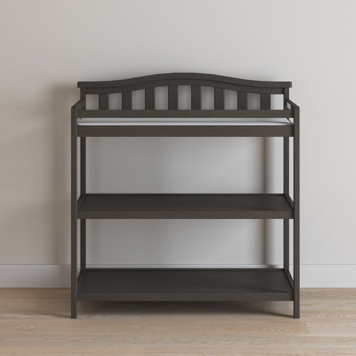 The Child Craft Arch Top Baby Changing Table in Dapper Gray features three open shelves, a slatted backrest, and raised sides for safety, offering stylish and functional nursery storage against a light wall and hardwood floor.