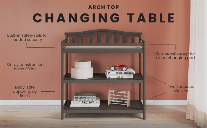 The Child Craft Arch Top Baby Changing Table in Dapper Gray features two spacious shelves, a changing pad, built-in safety rails, sturdy construction, and a baby-safe finish—perfect nursery storage by a peach wall and curtained window.