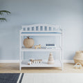 The Child Craft Arch Top Baby Changing Table in matte white, with a blue pad, wicker basket, wooden toys, books, and a stacking toy, sits against a light blue wall, paired with a striped rug, plant, and charming nursery decor.
