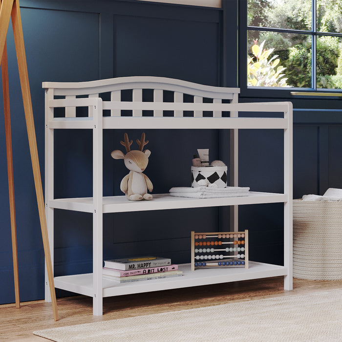 The Child Craft Arch Top Baby Changing Table in matte white features two shelves holding a plush deer toy, folded towels, a small box, and an abacus. Set by a sunlit window with greenery views against a blue paneled wall.