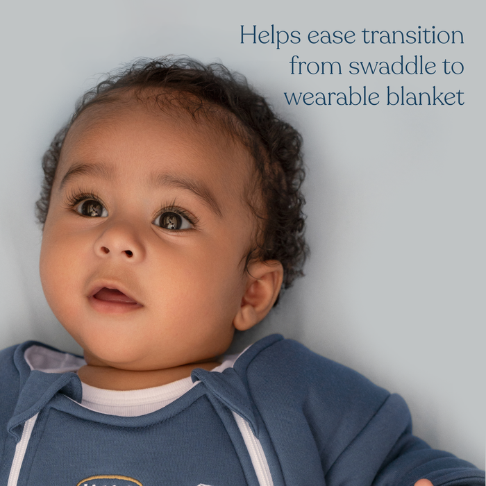 A curly-haired baby in blue lies on a soft surface. Text: HALO Baby Merlin's Magic Sleepsuit® 100% Cotton helps your baby transition from swaddle to a comfortable, wearable blanket.