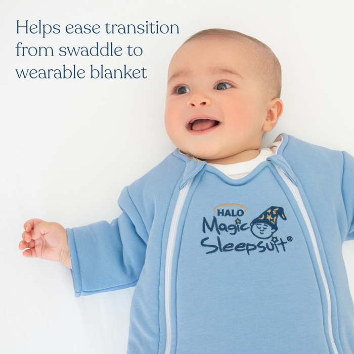 A smiling baby on a white surface wears the HALO Baby Merlin’s Magic Sleepsuit® 100% Cotton, designed to ease swaddle transition and support restful sleep during regressions.