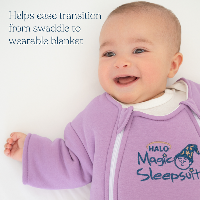 A smiling baby in a lavender Baby Merlin's Magic Sleepsuit® 100% Cotton by HALO lies on a white surface. Text reads: Eases swaddle transition and supports better sleep for babies.