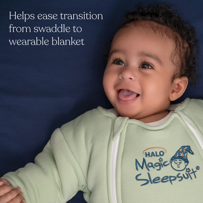A smiling baby in a light green Baby Merlin's Magic Sleepsuit® 100% Cotton by HALO lies on a dark blue surface. Text reads: Helps ease swaddle transition to wearable blanket. The sleepsuit features the HALO logo.