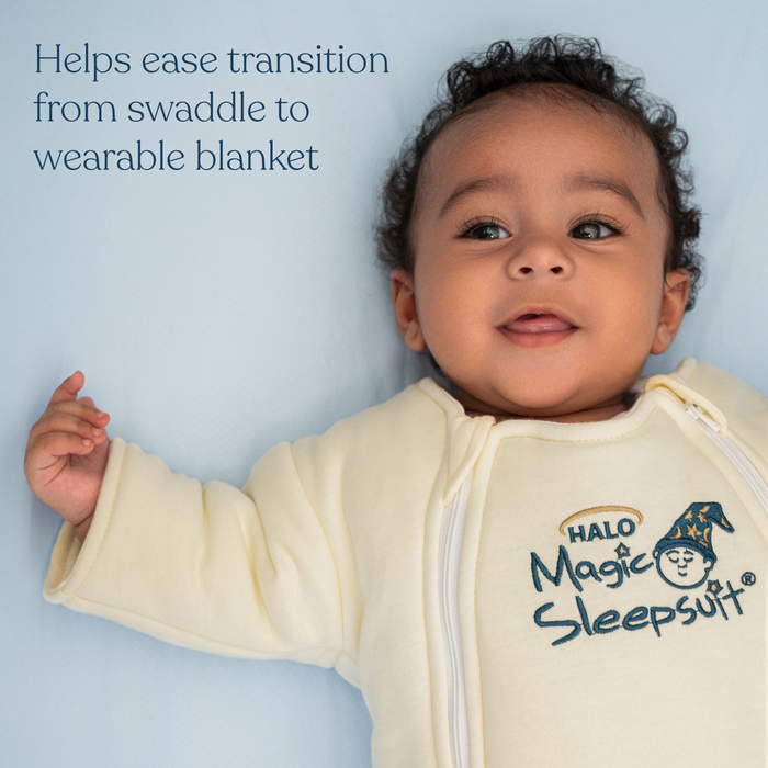 A smiling baby lies on a light blue surface wearing the HALO Baby Merlin's Magic Sleepsuit® 100% Cotton. Text beside the baby reads, Helps ease transition from swaddle to wearable blanket for a smoother swaddle transition.