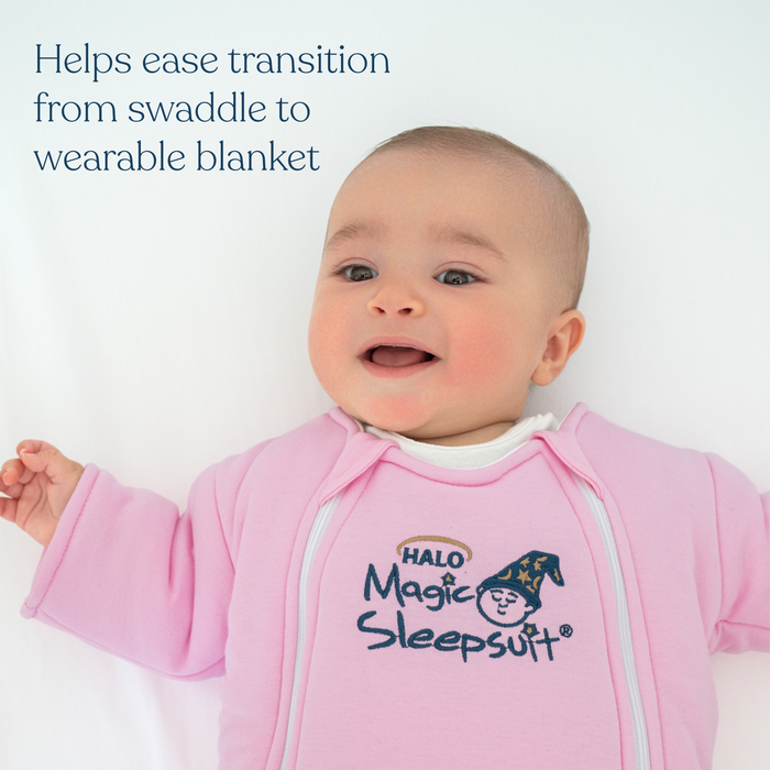 A smiling baby lies on a white surface, wearing a pink HALO Baby Merlin's Magic Sleepsuit® 100% Cotton. Blue text reads, Helps ease transition from swaddle to wearable blanket—a cozy innovation in baby sleepwear.