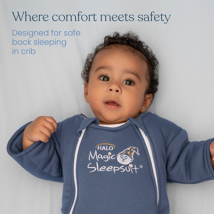 A baby in a blue HALO Baby Merlin's Magic Sleepsuit® 100% Cotton rests on a light blue surface, gazing upward. Caption: Comfort meets safety—ideal for swaddle transition and safe back sleeping in the crib.