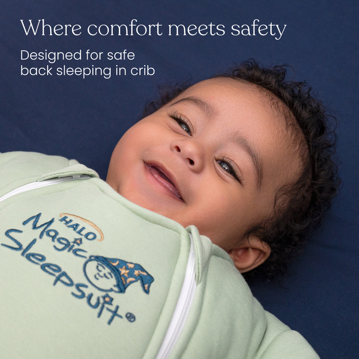 A smiling baby lies on a dark blue surface, wearing the HALO Baby Merlin's Magic Sleepsuit® 100% Cotton with a wizard hat logo. White text above reads: Where comfort meets safety. The perfect swaddle transition for safe back sleeping in crib.