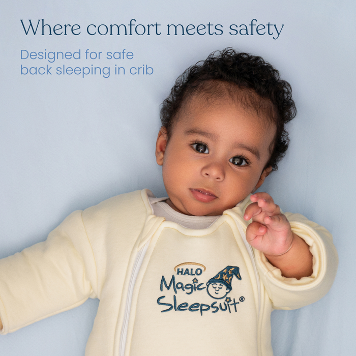 A baby with curly hair wears a light yellow Baby Merlin’s Magic Sleepsuit® 100% Cotton by HALO, lying on a pale blue sheet. Text: Where comfort meets safety. Designed for safe crib back sleeping—ideal as a swaddle transition suit.