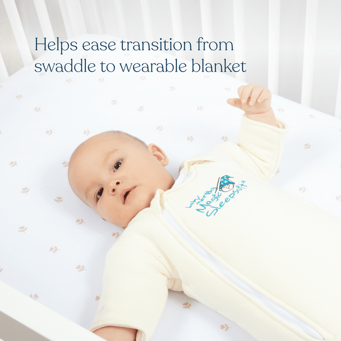 A baby lies in a crib on a white sheet with pink patterns, wearing the HALO Baby Merlins Magic Sleepsuit® 100% Cotton. Text above reads: Helps ease swaddle transition from swaddle to wearable blanket.
