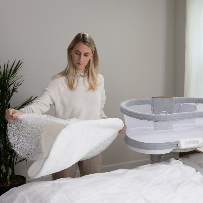 A woman stands beside a gray bassinet, lifting the HALO BassiNest® DreamWeave Breathable Pad with hypoallergenic mesh, in a softly lit room with a plant in the background.