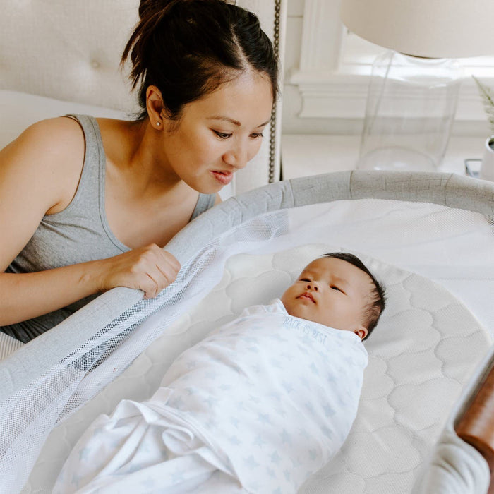 A woman smiles lovingly at a baby swaddled in white, resting on the HALO BassiNest® DreamWeave Breathable Pad in the bassinet. The softly lit bedroom creates a calm, nurturing atmosphere.