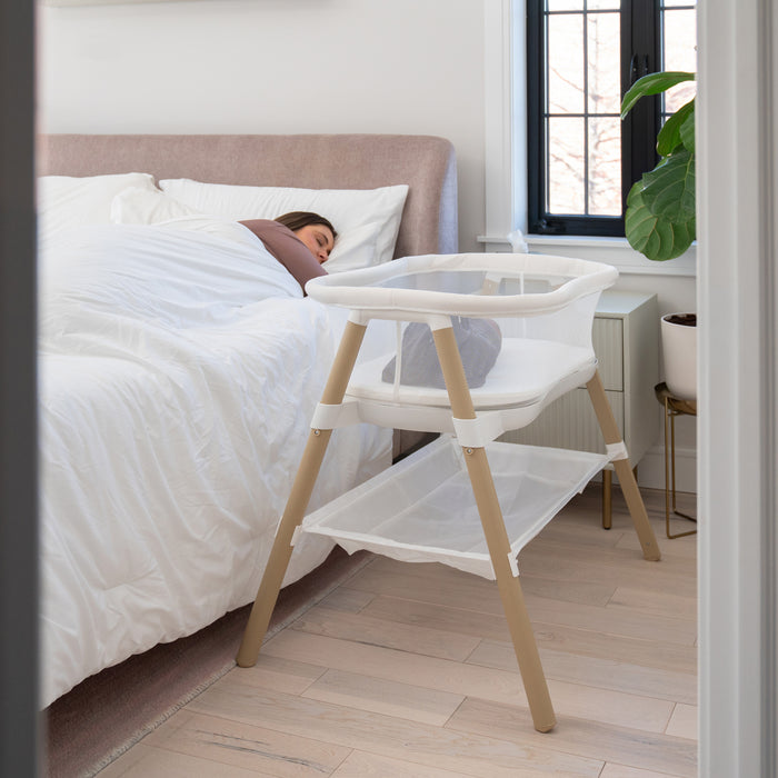 A woman sleeps in bed next to the HALO BassiNest® Essential Sleeper, a modern space-saving bassinet that's empty. The room has light wood floors, a plant, and a window with black frames.