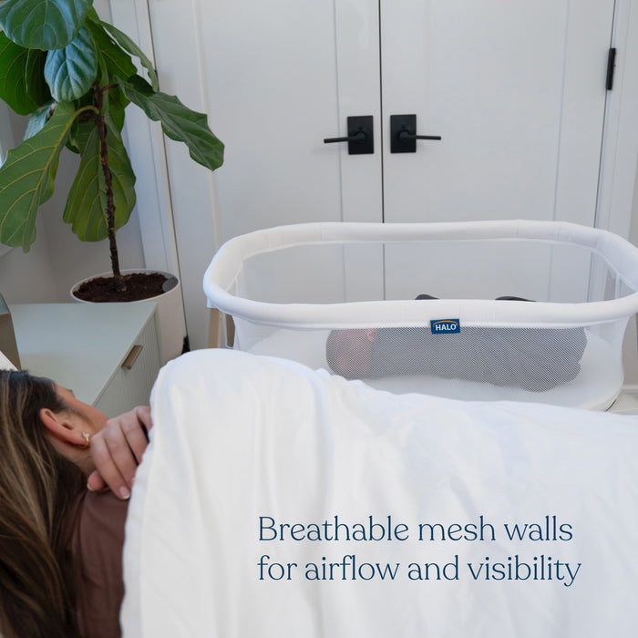 A woman sleeps beside the HALO BassiNest® Essential Sleeper, a bassinet featuring mesh walls for airflow and clear visibility of the baby. A green plant adds a touch of nature to this serene scene.