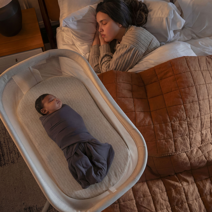 A woman sleeps in bed under a brown quilt while her swaddled baby rests in a bassinet with soft HALO BassiNest® Fitted Sheet 100% Organic Cotton 2-Pack, creating a cozy, serene bedroom atmosphere with warm lighting.