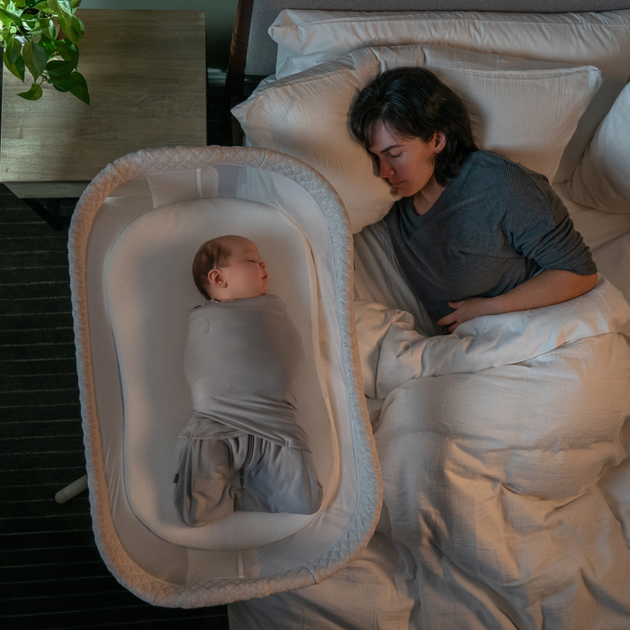 A woman sleeps in bed next to a baby swaddled in a bassinet with HALO BassiNest® Fitted Sheet 100% Organic Cotton Muslin 2-Pack. A green plant sits on the softly lit bedside table; both look peaceful and comfortable.