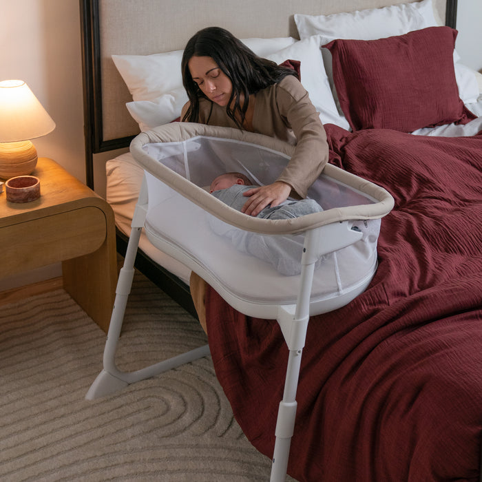 A woman gently soothes her baby lying in a HALO BassiNest® Lite Portable Bassinet beside a bed with burgundy bedding, while a lamp and candle on the nightstand add to the cozy, warm atmosphere.