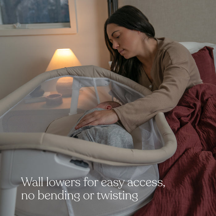 A woman gently touches a baby sleeping in a HALO BassiNest® Lite Portable Bassinet. The breathable mesh walls are lowered for easy access without bending or twisting. A lamp on the bedside table softly lights the serene room.