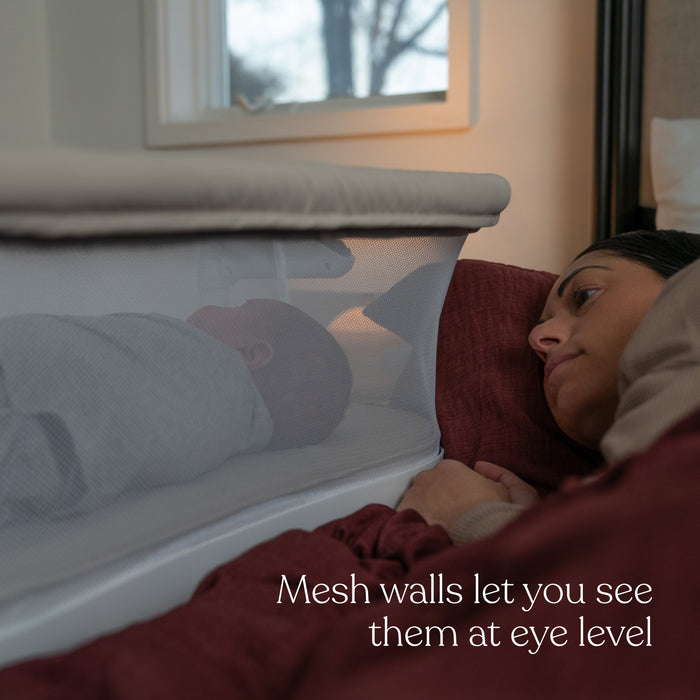 A woman rests in bed, watching a baby asleep in a HALO BassiNest® Lite Portable Bassinet featuring breathable mesh walls. Text reads, Mesh walls let you see them at eye level.