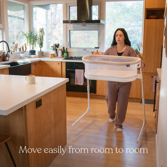 In a spacious kitchen with wooden cabinets and large windows, someone in cozy brown loungewear carries the HALO BassiNest® Lite Portable Bassinet, featuring breathable mesh walls. Text reads, Move easily from room to room.