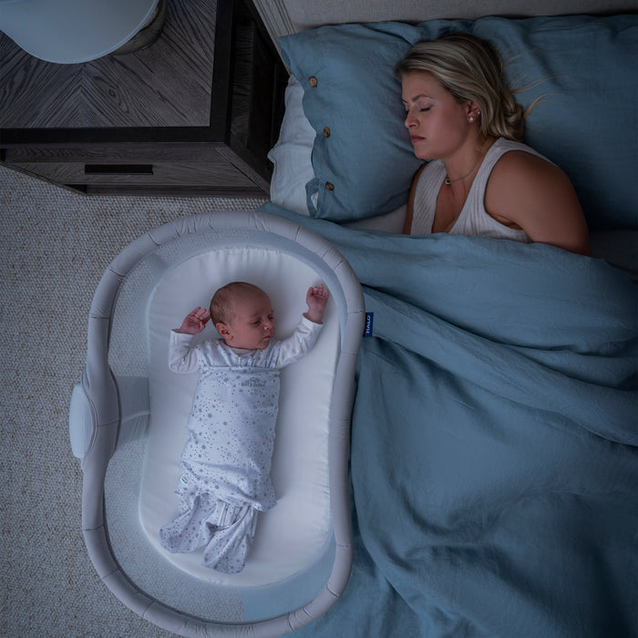 A woman sleeps in bed under a blue blanket while her baby rests beside her in the HALO BassiNest® Soothing Swivel Sleeper 3.0 Bassinet with breathable mesh walls, both viewed from above in a softly lit, cozy room.
