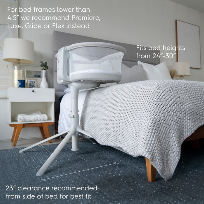 A bedroom features the HALO BassiNest Swivel Sleeper 3.0 next to a bed, accommodating heights from 24–30 with a 23 side clearance, ideal for versatility. A nightstand with a lamp and plant is in the background, all in soft, neutral tones.