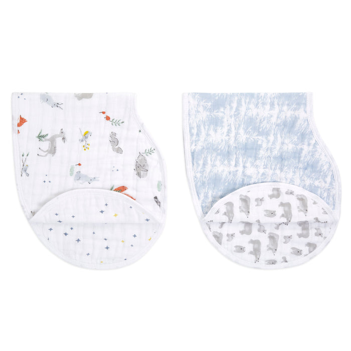 Two aden + anais boutique 100% cotton muslin burpy bibs are illustrated: the left features animal prints with colorful accents on absorbent muslin, while the right showcases a blue tie-dye pattern with gray bear silhouettes beneath. Both have curved edges and a luxurious finish.