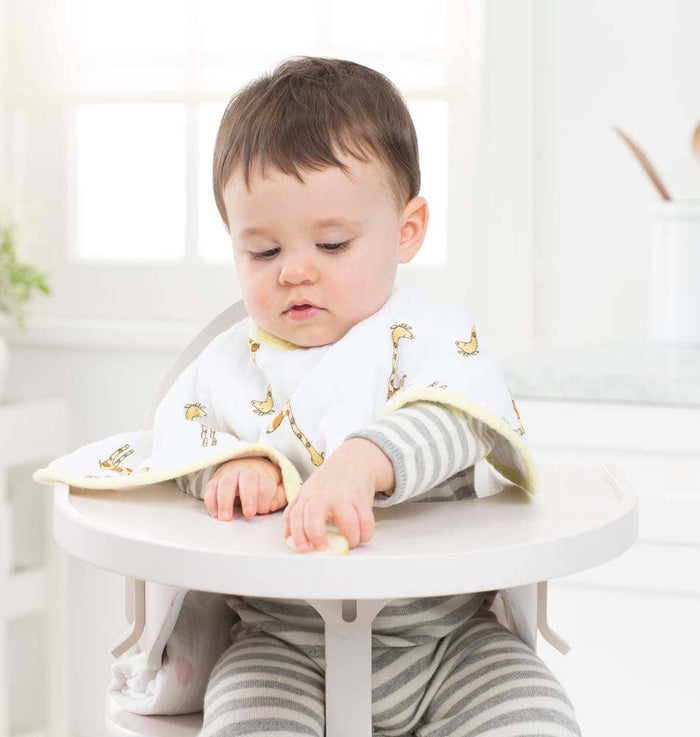 Wearing a giraffe-pattern aden + anais muslin burpy bib, a baby in a gray and white striped outfit sits in a high chair reaching for food. Sunlight streams through the window of the bright kitchen, illuminating the cozy scene.