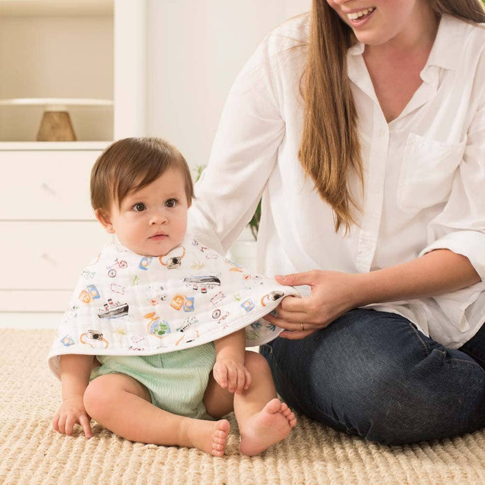 A baby with an aden + anais boutique 100% cotton muslin burpy bib featuring colorful patterns sits on a carpet beside a smiling woman in a white shirt and jeans. The softly lit room has a white chest and background items, enhancing the scenes luxurious finish.
