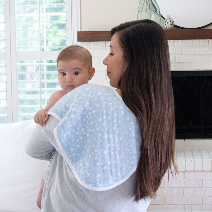 A woman cradles a baby over her shoulder, with an aden + anais boutique 100% cotton muslin burpy bib draped over it. Theyre in a room with white walls and a cozy fireplace, as sunlight filters through the window blinds.