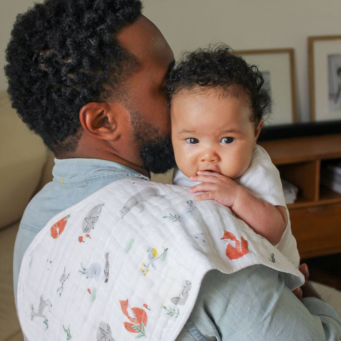 A man holds a baby resting on his shoulder, wrapped in an aden + anais boutique 100% cotton muslin burpy bib featuring colorful animal prints. The baby gazes thoughtfully at the camera, touching its lips, with a blurred background of frames and furniture adding a cozy ambiance.