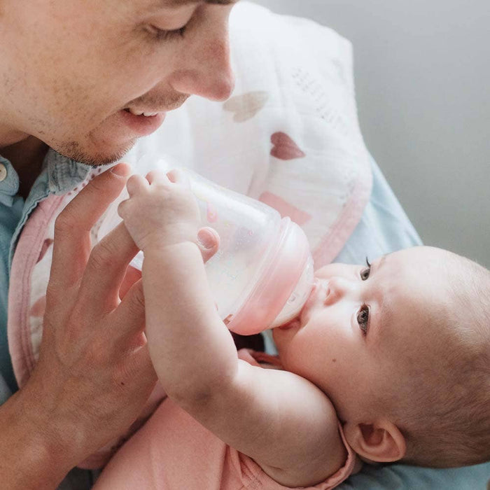 A person gently bottle-feeding a baby wrapped in a pink blanket, using aden + anais boutique 100% cotton muslin burpy bibs to keep everything tidy. The scene conveys a sense of warmth and care.
