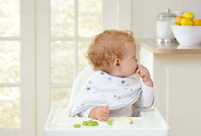 A curly-haired baby sits in a high chair, wearing an aden + anais boutique 100% cotton muslin burpy bib with star patterns. The absorbent bib catches any spills as the baby holds food, and grapes and an apple slice rest on the tray. A bowl of lemons is visible in the kitchen background.