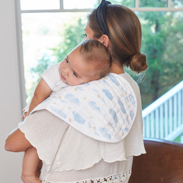 A woman wearing a headband gently holds a baby on her shoulder, using an aden + anais boutique 100% cotton muslin burpy bib. Standing by a window with lush greenery outside, she wears a white blouse with lace detailing that complements the soft muslin of the bib.