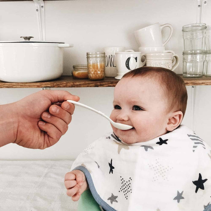 A smiling baby wears an aden + anais boutique 100% cotton muslin burpy bib, adorned with stars, while being fed by an adult. The absorbent design ensures a mess-free mealtime. In the background, kitchen shelves display dishes, jars, and cups.