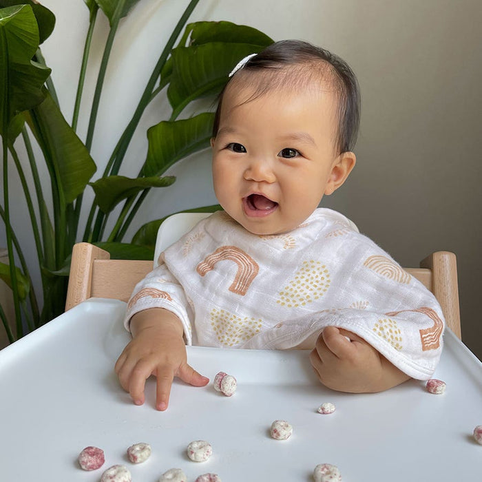 A smiling baby in a high chair reaches for snacks on the tray, wearing HALOs cheerful Tried & True Bundle Burpy Bib with rainbows. Large green leaves in the background add a natural touch to this joyful scene.