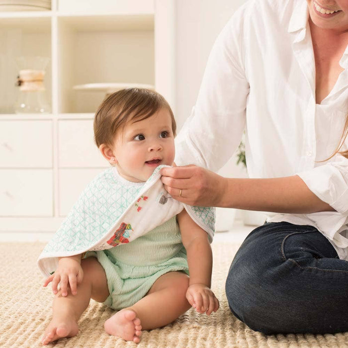 A baby in a light green outfit sits on a carpet, smiling as an adult kneels beside them, using an aden + anais boutique 100% cotton muslin burpy bib to gently wipe the babys mouth. A white bookshelf and soft lighting in the background enhance the cozy atmosphere.