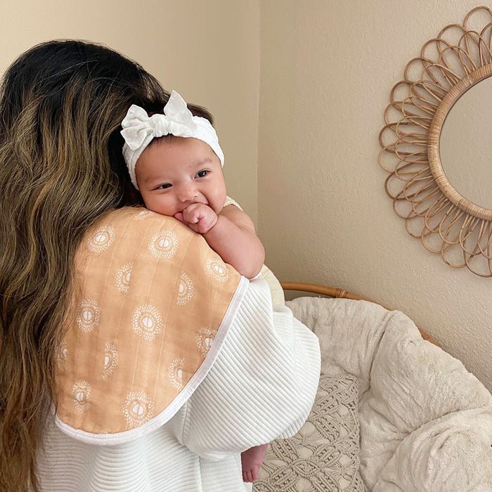 A woman with long hair cradles a baby on her shoulder, who wears a white headband with a bow. The baby rests comfortably against an absorbent aden + anais boutique 100% cotton muslin burpy bib, while behind them a decorative mirror enhances the warmth of the scene.