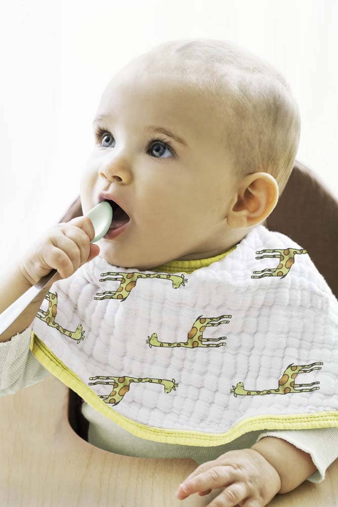 A light-skinned baby sits in a high chair, wearing an adorable giraffe-themed bib from the aden + anais boutique 100% cotton muslin burpy bibs 2 pack, holding a spoon near their mouth and looking upwards, creating a delightful scene against a bright, neutral background.