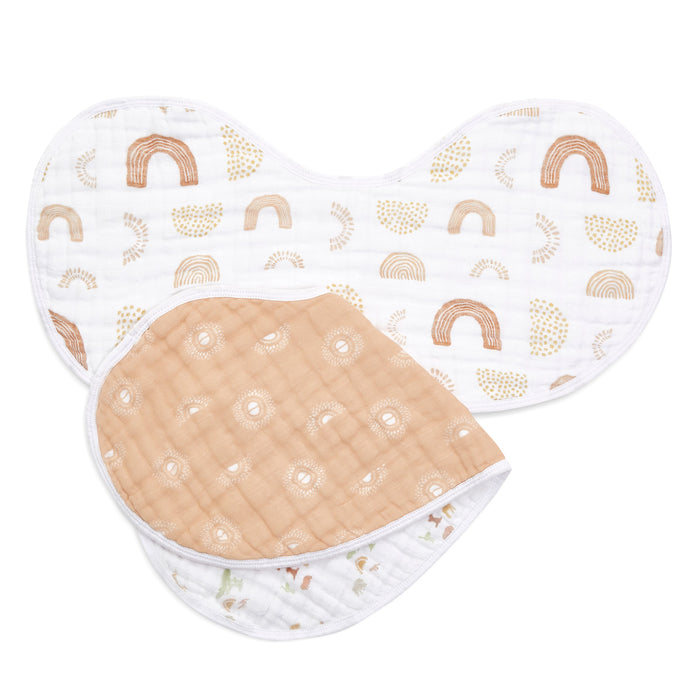 Two aden + anais boutique 100% cotton muslin burpy bibs are shown; one has a white background with orange rainbows, and the other has a beige side with white suns and a reverse of white with small animal prints. These absorbent layered cloths are arranged to overlap.