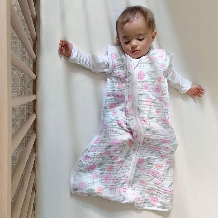 A baby sleeps peacefully in a crib, wearing an aden + anais boutique 100% cotton muslin light sleep sack TOG 1.0, featuring pink floral designs. The baby also wears a white long-sleeve shirt beneath the sleep sack, surrounded by the cribs light wooden bars.