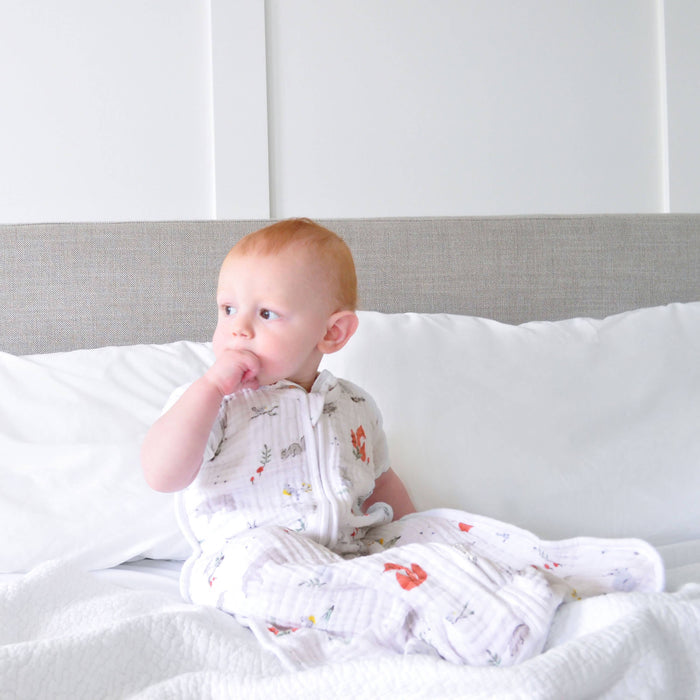 A baby with light hair sits on a bed, against a white pillow and gray headboard, wearing the aden + anais boutique 100% cotton muslin sleep sack TOG 1.0 in colorful patterns. The baby looks to the side, with one hand touching their mouth.