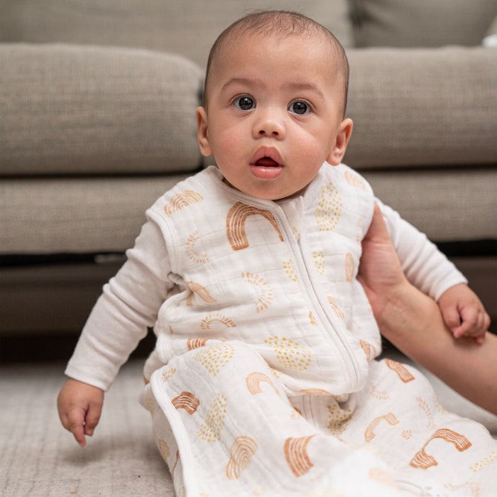 A baby sits on the floor in an aden + anais boutique 100% cotton muslin light sleep sack TOG 1.0, with an adults hands supporting them and a beige sofa elegantly in the background.
