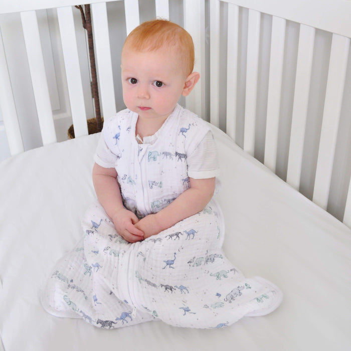 A small child with red hair sits on a white crib mattress, wrapped in an aden + anais boutique 100% cotton muslin light sleep sack with animal prints (TOG 1.0). The child looks forward calmly while the crib features slatted sides.