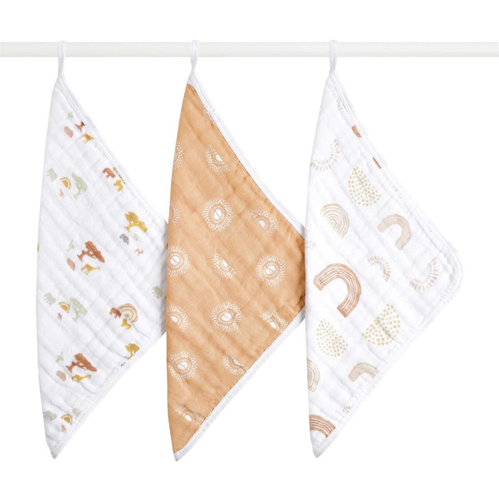 Three hanging aden + anais boutique 100% cotton muslin oversized washcloths: one with small animal illustrations on thicker fabric, another featuring white circular designs on a brown base, and the third displaying abstract shapes in soft colors.