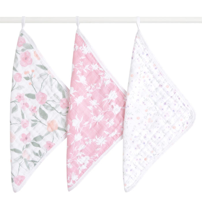 Three aden + anais boutique 100% cotton muslin oversized washcloths hang from a rod: one with a pink and green floral pattern ideal for delicate skin, another featuring pink with white floral designs, and the third is white adorned with small pastel dots, all ensuring comfort during tender moments.