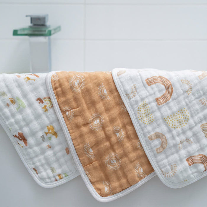 Three aden + anais boutique 100% cotton muslin oversized washcloths drape over a bathtub edge, showcasing patterns of animals, a central brown and white design, and rainbows. The bathroom background is white and minimalistic.